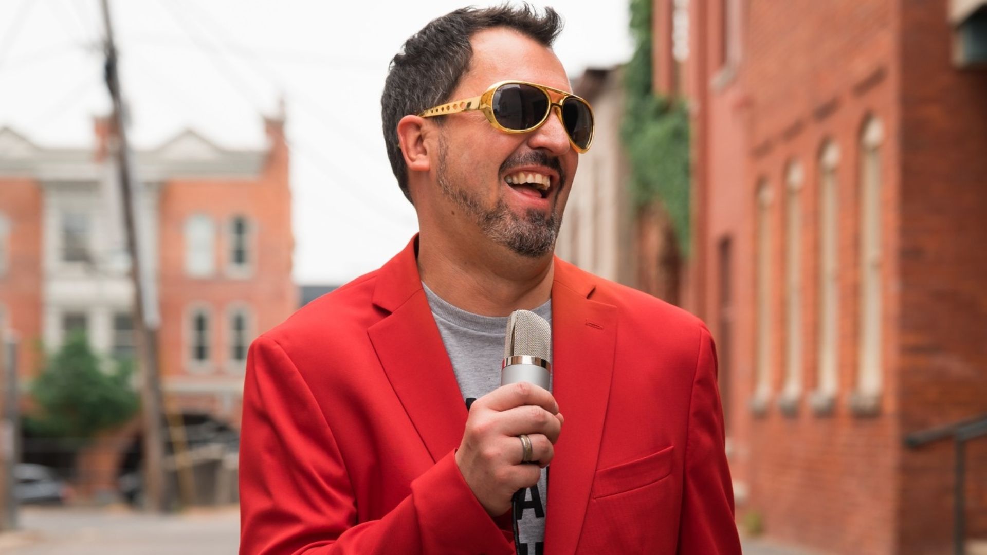 A man in a jacket and shades holding a microphone outdoors.